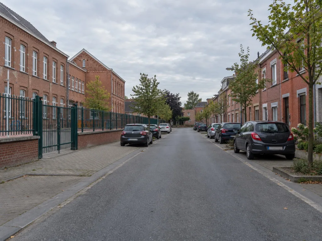 Rustige straat langs een bakstenen schoolgebouw met een gesloten hek, geparkeerde auto's aan beide kanten en geen kinderen in zicht.